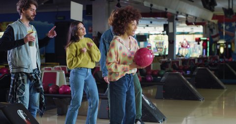 Cinematic shot of happy young african female bowler throwing strike ball and exulting victory while having fun to play bowling together with multiracial college friends on weekend in sporting club.