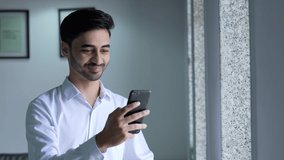 Young Indian man holding smart phone using mobile apps watching funny video. Smiling man having fun chatting in social media. Asian male chatting with friend on Smart Phone background office.