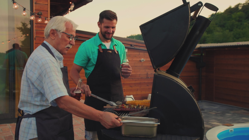 Senior man barbecuing with his son in the backyard. Both of them wearing aprons. Gray haired man holding bottle of beer while his son controlling roasting meat and holding barbecue gripper | Shutterstock HD Video #1087543976