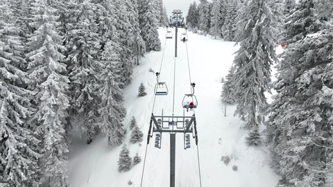 Aerial view over snowy mountain winter forest with chair lift at ski resort. Ski lift cable car top down drone shot. Winter landscape scene with snow covered trees. Outdoor tourism skiing snowboarding