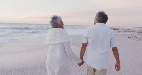 Back view of happy hispanic just married senior couple walking on beach at sunset. love, romance and senior marriage concept.