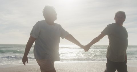Happy hispanic senior couple holding hands, walking on beach at sunset. love, romance and active retirement lifestyle.