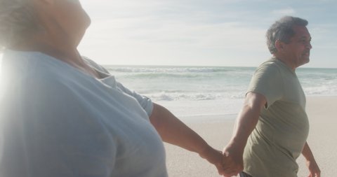 Happy hispanic senior couple holding hands, walking on beach at sunset. love, romance and active retirement lifestyle.