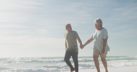 Happy hispanic senior couple holding hands, walking on beach at sunset. love, romance and retirement lifestyle.