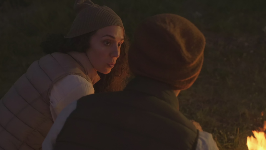 Horizontal shot of man and woman in love spending evening in camp on lakeside sitting close to campfire chatting about something