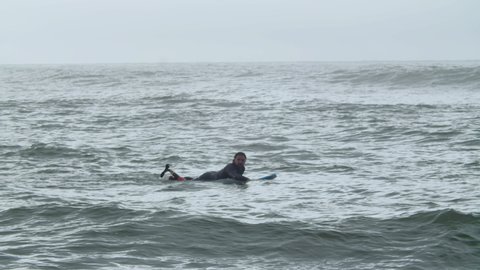 Sportsman in wetsuit lying on surfboard in ocean. Long shot of fit surfer with artificial leg training, surfing, swimming, devoting time to physical activity. Disability, extreme sport concept
