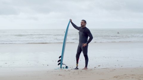 Cheerful surfer with artificial leg standing on sandy beach. Long shot of handsome man in wetsuit standing on seashore with surfboard, smiling at camera, showing shaka sign. Greeting, sport concept