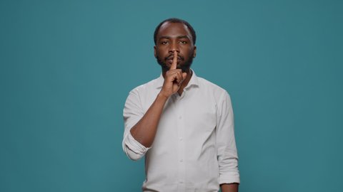Portrait of male model showing silence sign in front of camera, putting index finger over mouth to keep secret. Person advertising hush and mute gesture, privacy and confidential information.