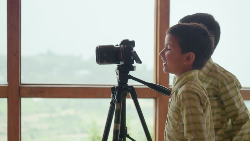 Two young happy Indian male school children are playing around a digital photography camera or DSLR indoors with a background of misty mountainous valley out of window. Concept of Practical education | Shutterstock HD Video #1087570541