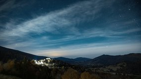 Night starry sky with white clouds floating above small town illuminated by street lights in highland near mountains 4K time-lapse