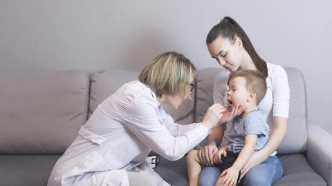 pediatrician doctor visits baby boy child at home. caucasian kid sitting on mother's knees grey sofa living-room. female woman doctor white uniform, examine child's throat, tongue, neck, lymph nodes 