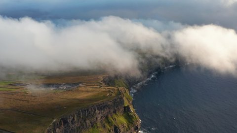 Aerial panoramic shot of high cliffs shrouded in low clouds. Romantic natural scenery of sea coast at golden hour. Cliffs of Moher, Ireland