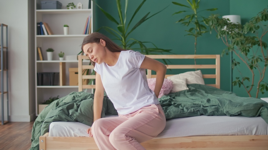 Young Woman Feels Back Pain on the Bed at Home, Rubs Her Lower Back, Trying To Relieve Muscle Tension. Uncomfortable Bed or Mattress, Poor Sleep, Stiffness in Movement, Worsening of Chronic Back Pain. Royalty-Free Stock Footage #1087578647