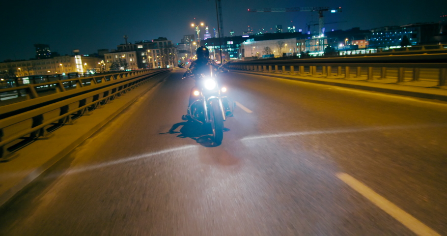 Unrecognizable Authentic Motorcyclist is Riding fast on City roads and streets at night with Warsaw Skyline at Horizon. Biker drives Motorbike in dark foggy evening with lights. 4K wide tracking shot