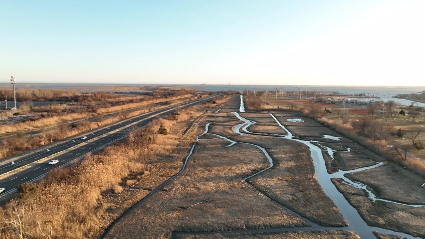 An aerial view over a marsh on Long Island, NY. A long road with a few cars run next to it, shot on a sunny day. The camera dolly in over the grass and water pathways.