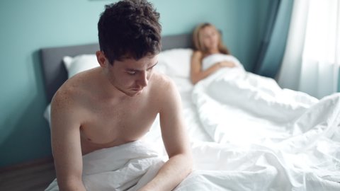 Frustrated man sitting on bed on different side of woman. Young unhappy couple having problem in bedroom, quarrel between lovers. Divorce, marriage, conflict, family crisis, love concept.