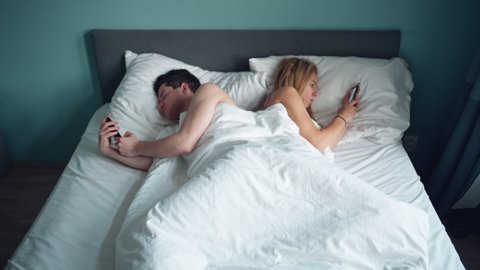 Married young multi-ethnic couple lying in the bed back to back at night and typing or scrolling on smartphones. Young unhappy couple problem in bedroom, quarrel between lovers. Divorce, marriage