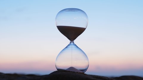 Hourglass time lapse shot, blurred evening sky on back. Quickly night falls, picture becomes dark. Slowly at first, and then faster and quite quickly, the remains of the sand from upper bulb 스톡 비디오