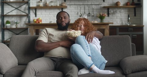 Young interracial family watching horror movie on TV with popcorn sitting on couch in apartment. Scared couple watching terrible videos together on tv at home