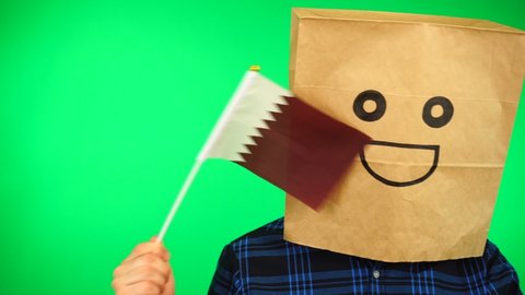 Portrait of man with paper bag on head waving Qatari flag with smiling face against green background.