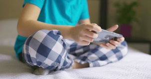 Kid is using smartphone, child browsing internet, talking on smart phone, boy communicating with parents or friends while sittng on a bed