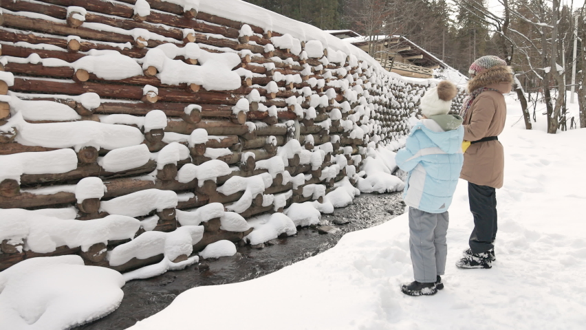 Side view of two girls in outerwear standing in front of small cascade waterfall in snowy forest. Female kids looking at water stream flowing down with wooden dam on background. Concept of landscape.
