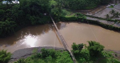 Drone shot of A wooden suspension bridge built over a large river.  Beside the river there are rice fields. The water flowing in the river is brown. The bridge was named "Jembatan Gantung Kali Progo"