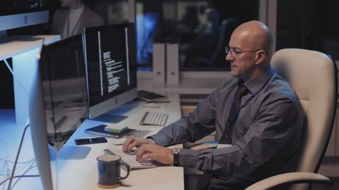 Side view of mature Caucasian male cyber security specialist wearing eyeglasses and formal attire, sitting at desk in office at night, coding on PC