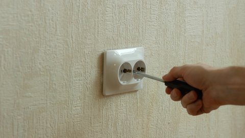Close-up of a man repairing an electrical outlet in an apartment. An electrician fixes a loose outlet protruding from the wall with a screwdriver. Electric installation work.