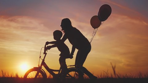 mom teaches son to ride a bike. happy family kid dream concept. mom and child son learn to ride a bike silhouette in the park in nature. sunlight happy family doing sports outdoors in the park .