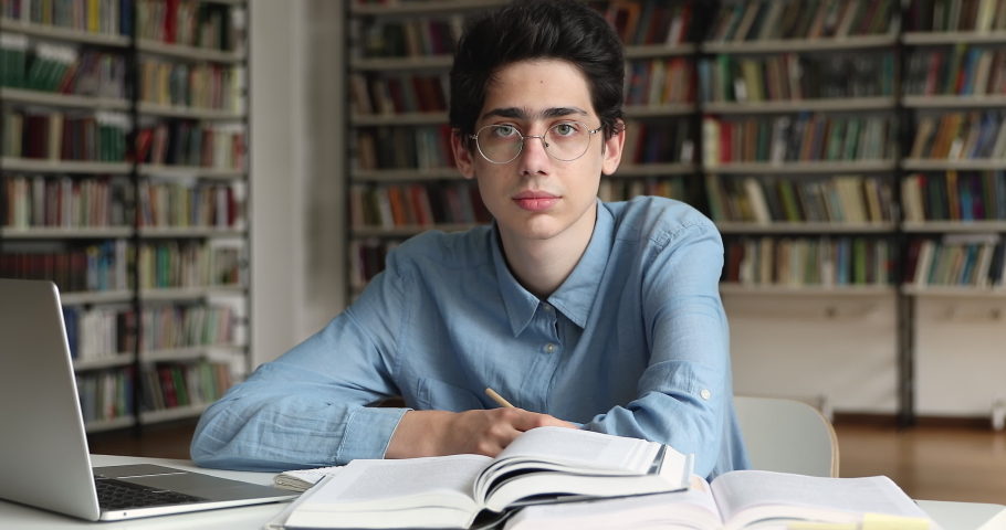 Portrait clever young guy in glasses teenage high schooler pose at work desk at library distracted from pc books. Smart gen z man college university student look at camera start smiling enjoy learning Royalty-Free Stock Footage #1087646105