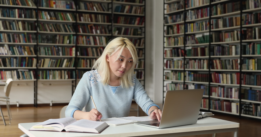 Busy young Asian woman study on distance take notes of digital lecture video lesson internet course sitting at library desk by laptop. Serious teenage lady undergraduate write essay using online info Royalty-Free Stock Footage #1087646171
