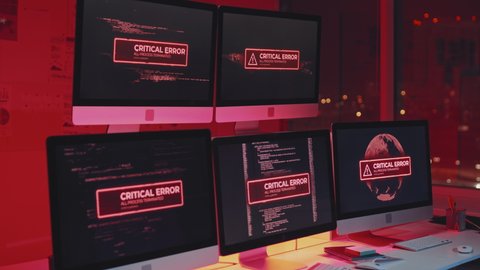 Zooming in of critical error warning message showing on multiple computer screens on desk in office at night, red light flashing