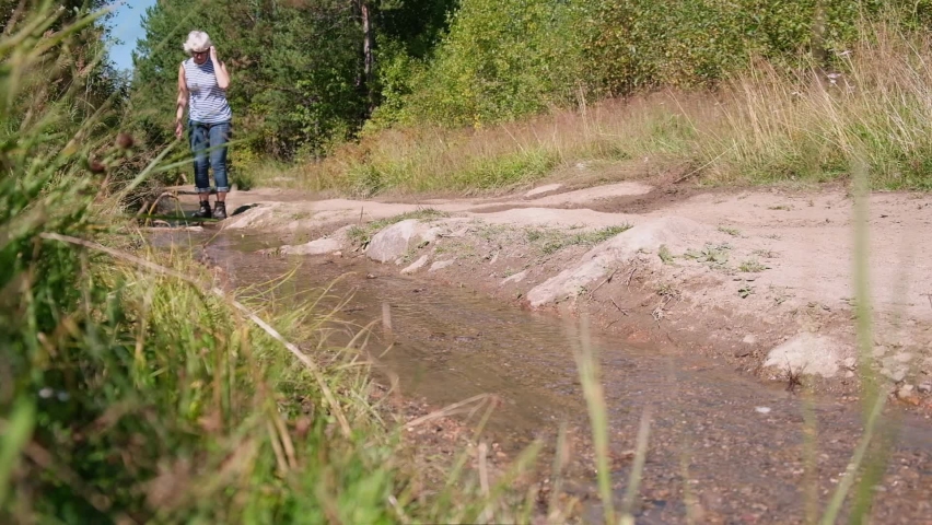 Adult female traveler in jeans and trekking boots walks along forest road with stream.Close-up of female legs walking in slow motion.Feet in hiking boots are walking through the woods through a stream