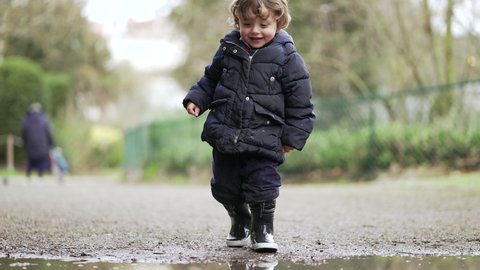 Little baby boy jumping into water puddle and running in mud water wearing boot rains