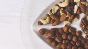 Various nuts close-up on a white plate on a white wooden table. View from above. 4k raw video with smooth camera movement.