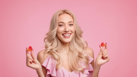 Smiling Blonde Lady Holding Two Heart Shaped Jelly Candies Posing Over Pink Studio Background. Love And Romantic Relationship. Sweet Tooth Concept. Slow Motion