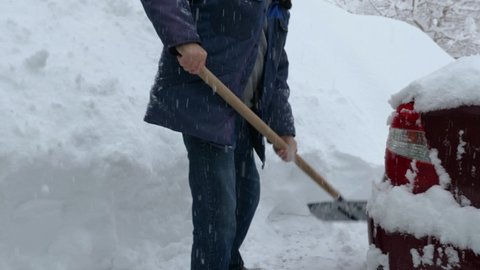 Timelapse of a man in a cap, jacket, and jeans shoveling snow on a huge snowdrift. Snow falls from the sky