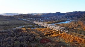 Railroad bridge over river in highland valley with colorful trees and bushes and speeding train under blue sky on sunny autumn day aerial drone 4K