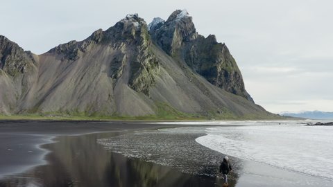 Drone Shot Of Man On Black Sand Beach Walking As The Tide Comes From Vestrahorn Mountain, Iceland