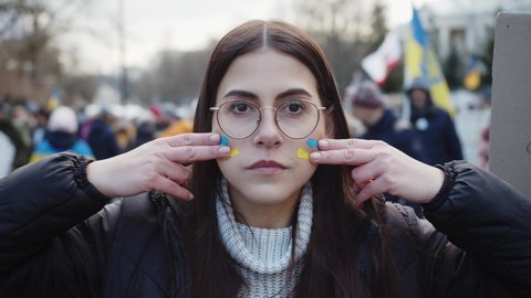 WARSAW, POLAND - FEBRUARY 27 2022: Girl Paints The Flag Of Ukraine With Fingers On Face, Stands On Demonstration, Protest Against The Russian War Invasion In Ukraine. Ukrainian Flag, Protesters