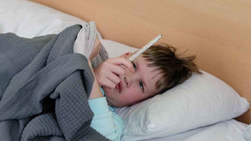 A sad boy is lying on the bed and measuring the temperature with a mercury thermometer, and he has a headache. The face is distorted with pain | Shutterstock HD Video #1087669016