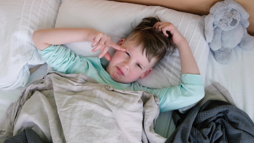 A sad boy lies on the bed with a fever and headache and rubs his head in pain. The face is twisted with pain | Shutterstock HD Video #1087669031