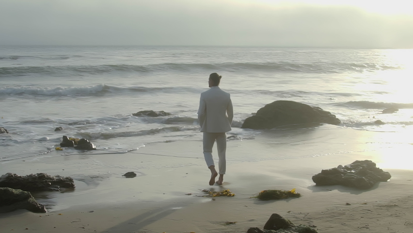 A young male model in a white suit walks by the ocean on the shore Royalty-Free Stock Footage #1087670267