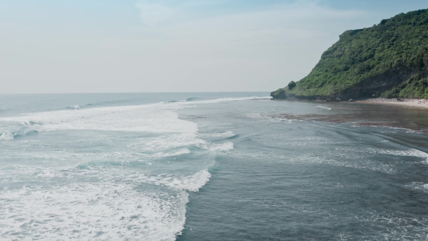 Aerial flyby of large ocean waves following each other and breaking into white foam on stone reef at high tide. Coast of tropical island with white sand and large rocks. Bali island drone view. | Shutterstock HD Video #1087692911