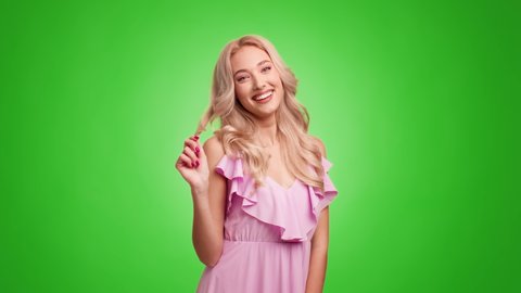 Gorgeous Blonde Lady Playing With Hair And Blowing A Kiss Smiling To Camera Standing Over Green Studio Background, Wearing Pink Dress. Female Beauty, Haircare Concept. Slow Motion