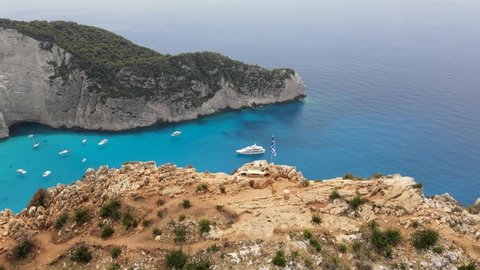 Amazing Navagio Beach with shipwreck on Zakynthos, Ionian Islands, Greece. Greek flag waving in the wind. Panoramic Aerial View 4K