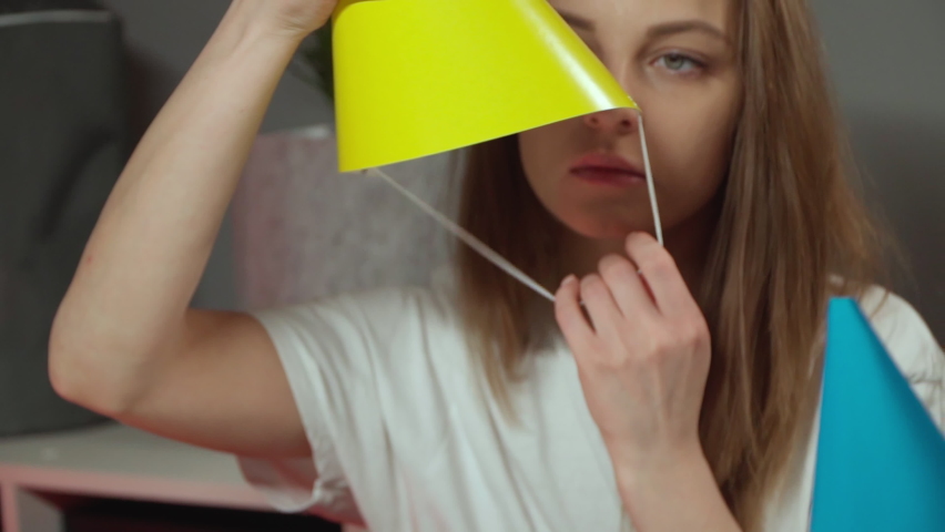Caucasian girl sits in a festive cap and smiles. A European woman in a white T-shirt puts on a birthday cap Decor for the holiday with your own hands. Homemade attributes for a birthday. Gray walls