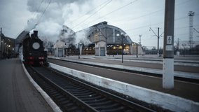 Steam locomotive train approaching station passing through a goods yard leaking smoke and steam ignited from behind, creating an atmospheric.