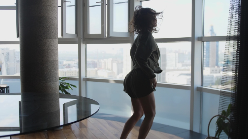 Happy carefree young woman in pajamas dancing alone in modern living room with a big window listen to music on smartphone.